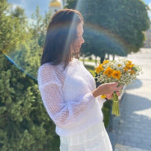May include: A woman in a white, lacy, knitted sweater and a white skirt holds a bouquet of yellow and white flowers.