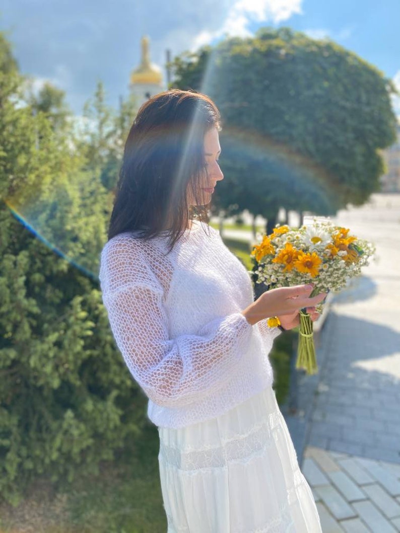 May include: A woman in a white, lacy, knitted sweater and a white skirt holds a bouquet of yellow and white flowers.