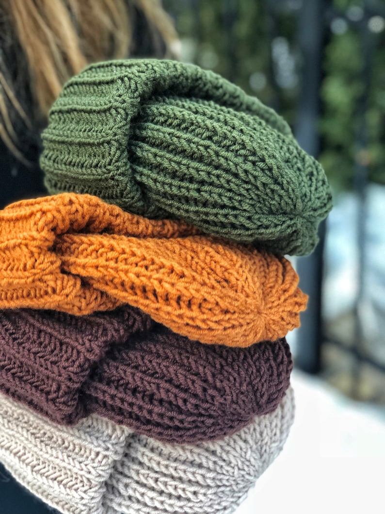 Puede incluir: Una pila de cuatro gorros de punto en diferentes colores: verde, naranja, marr&oacute;n y beige. Los gorros est&aacute;n doblados y apilados uno encima del otro.