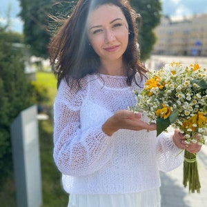 May include: A woman in a white crocheted sweater and a white skirt holds a bouquet of white and yellow flowers.