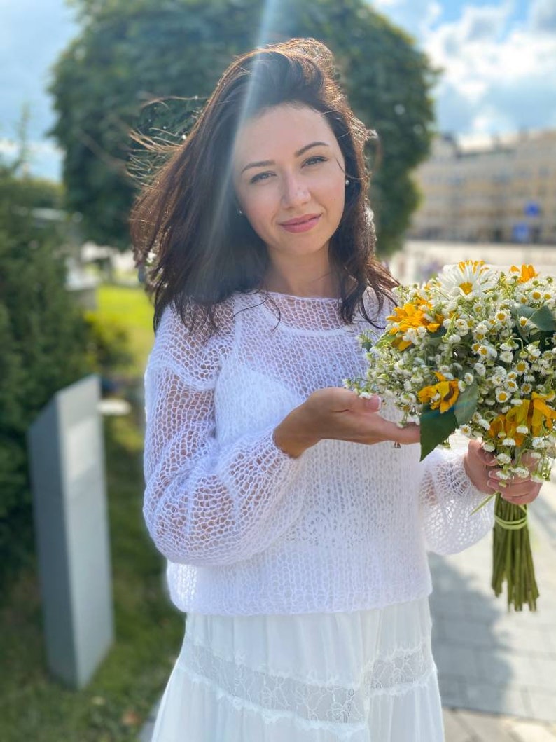 May include: A woman in a white crocheted sweater and a white skirt holds a bouquet of white and yellow flowers.