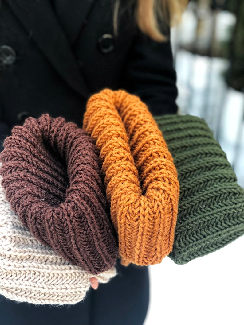 Puede incluir: Tres bufandas infinitas de punto en marr&oacute;n, naranja y verde. Las bufandas est&aacute;n dobladas y se sostienen en la mano de una persona.