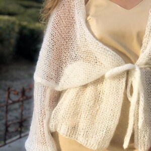 May include: A white, open knit cardigan with a tie closure. The cardigan is made of a lightweight, airy fabric and has a relaxed fit.