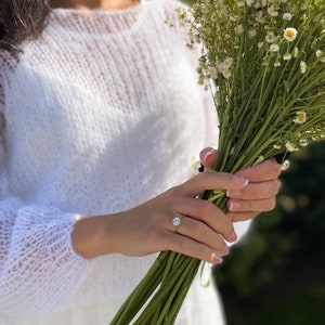 May include: A person wearing a white knitted sweater holds a bouquet of white flowers with yellow centers. The person is wearing a ring on their left ring finger.