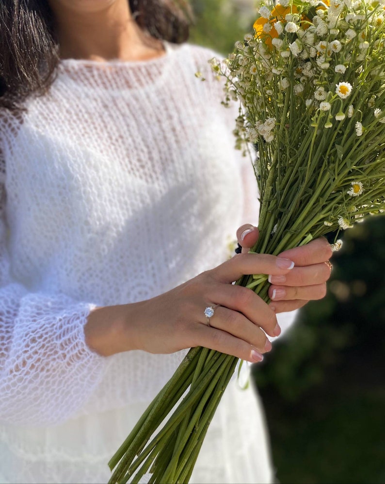 May include: A person wearing a white knitted sweater holds a bouquet of white flowers with yellow centers. The person is wearing a ring on their left ring finger.