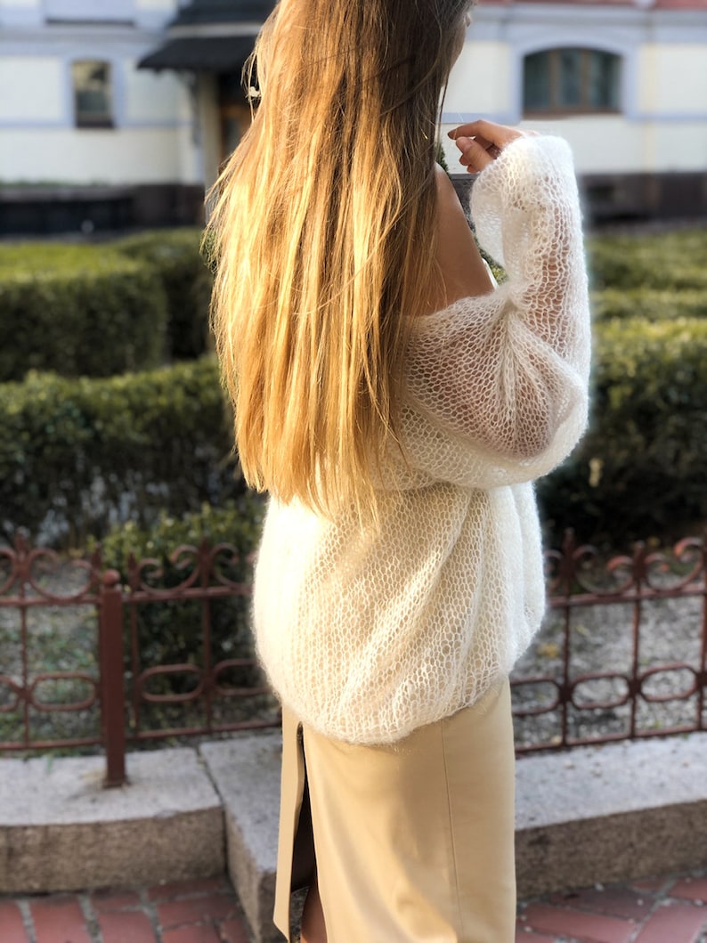 May include: A white, lightweight, knitted sweater with a loose fit and long sleeves. The sweater is made of a delicate, open-weave fabric.