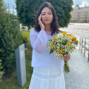 May include: A woman wearing a white knitted sweater and a white skirt with lace trim holds a bouquet of white and yellow flowers.