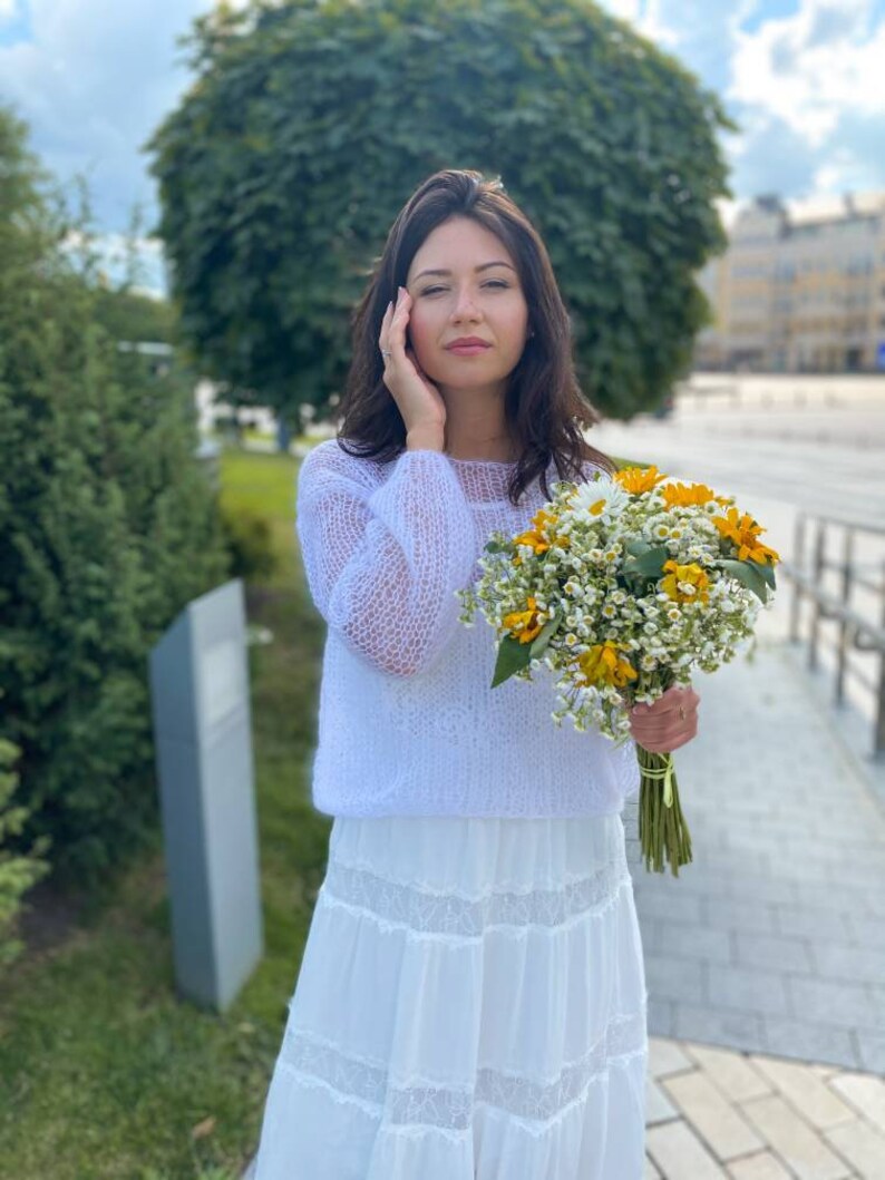 May include: A woman wearing a white knitted sweater and a white skirt with lace trim holds a bouquet of white and yellow flowers.