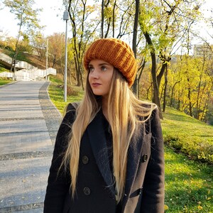 Puede incluir: Una mujer con un gorro de punto marr&oacute;n y un abrigo negro de doble botonadura. Est&aacute; de pie en un parque con &aacute;rboles en el fondo.