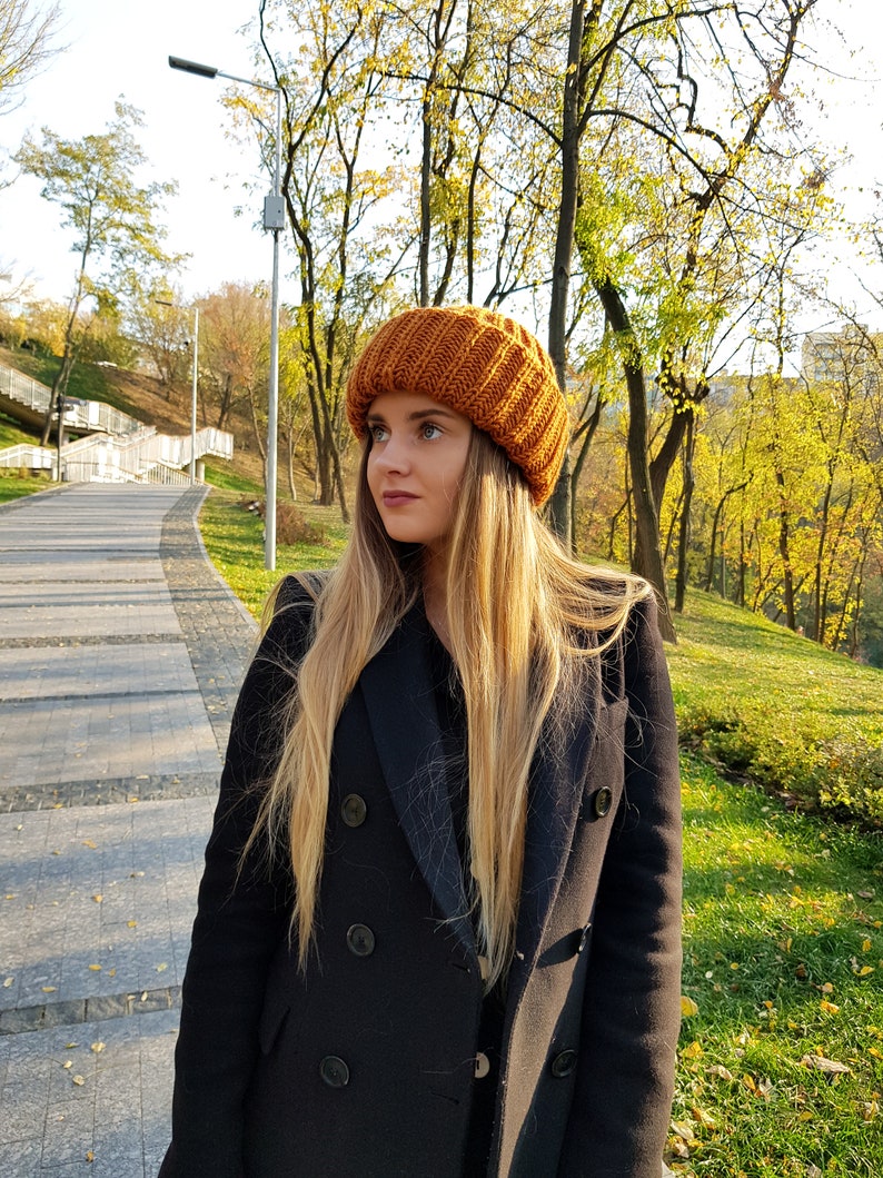 Puede incluir: Una mujer con un gorro de punto marr&oacute;n y un abrigo negro de doble botonadura. Est&aacute; de pie en un parque con &aacute;rboles en el fondo.