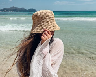 May include: A woman wearing a white sheer dress and a wide-brimmed straw hat stands on a sandy beach with the ocean in the background.