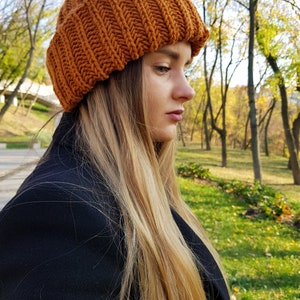 Puede incluir: Una mujer con el pelo largo y rubio lleva una boina de punto marr&oacute;n. La boina tiene una textura acanalada y est&aacute; bajada sobre su frente. Lleva un abrigo negro y est&aacute; de pie en un parque con &aacute;rboles en el fondo.