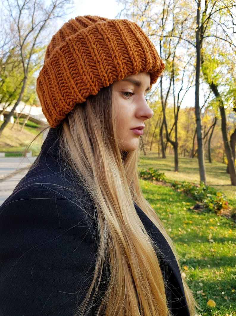 Puede incluir: Una mujer con el pelo largo y rubio lleva una boina de punto marr&oacute;n. La boina tiene una textura acanalada y est&aacute; bajada sobre su frente. Lleva un abrigo negro y est&aacute; de pie en un parque con &aacute;rboles en el fondo.