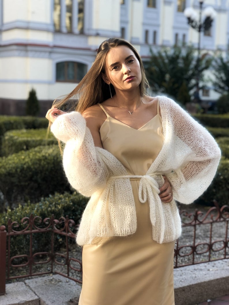 May include: A white knitted cardigan with a delicate open weave and long sleeves. The cardigan is tied at the waist with a matching sash. The model is wearing a beige slip dress underneath.