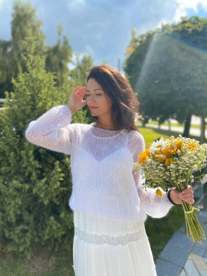 May include: A woman wearing a white, lacy, knitted sweater and a white skirt with lace trim. She is holding a bouquet of yellow and white flowers.