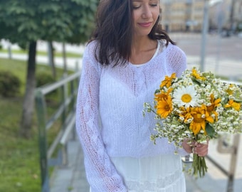 Può includere: Una donna indossa un maglione di lana bianco e una gonna bianca con pizzo e tiene un mazzo di fiori gialli e bianchi.