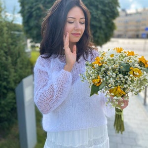 May include: A woman wearing a white crocheted sweater and a white skirt holds a bouquet of yellow and white flowers.