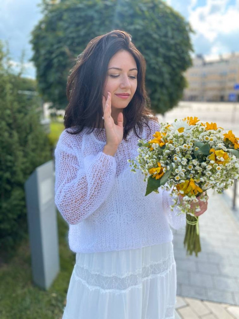 May include: A woman wearing a white crocheted sweater and a white skirt holds a bouquet of yellow and white flowers.