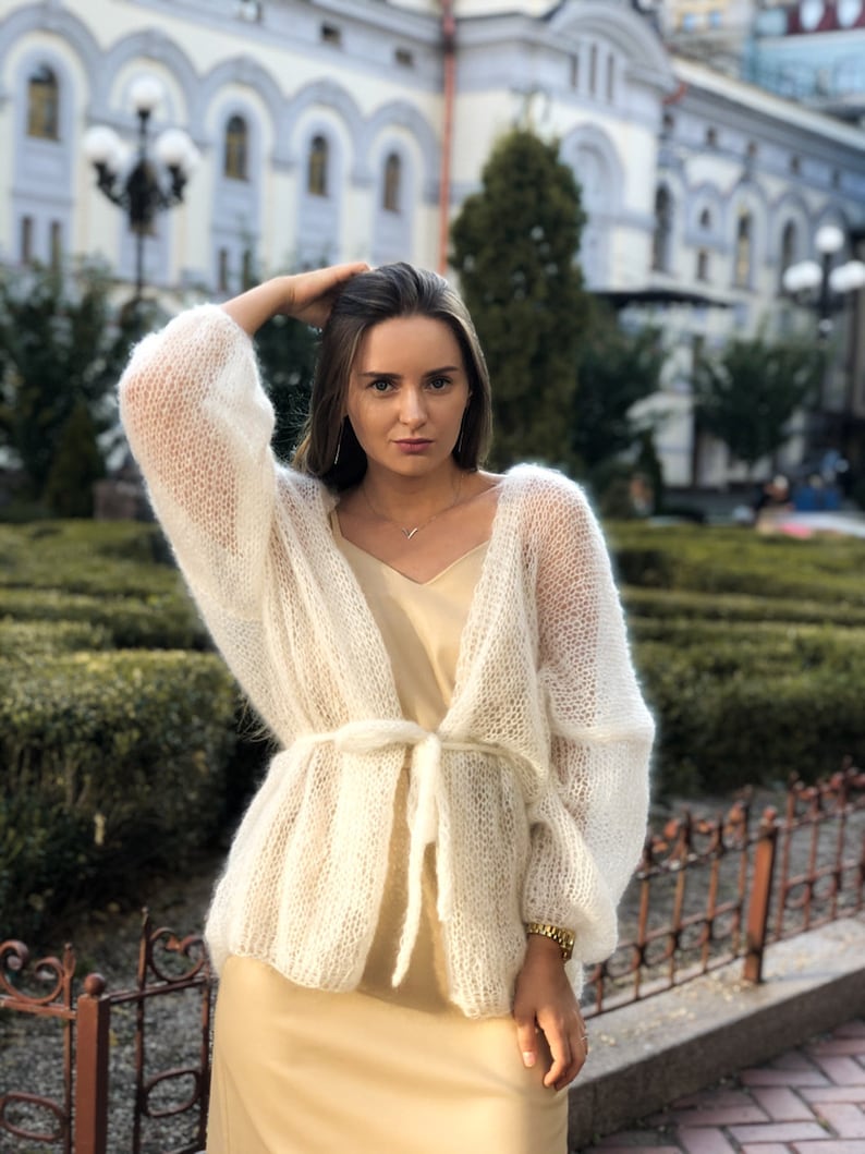 May include: A white, lightweight knit cardigan with a tie belt. The cardigan is made of a soft, airy yarn and has a relaxed fit.