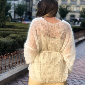May include: A white, sheer, knitted sweater with a loose fit and long sleeves. The sweater is tied at the waist with a white ribbon.