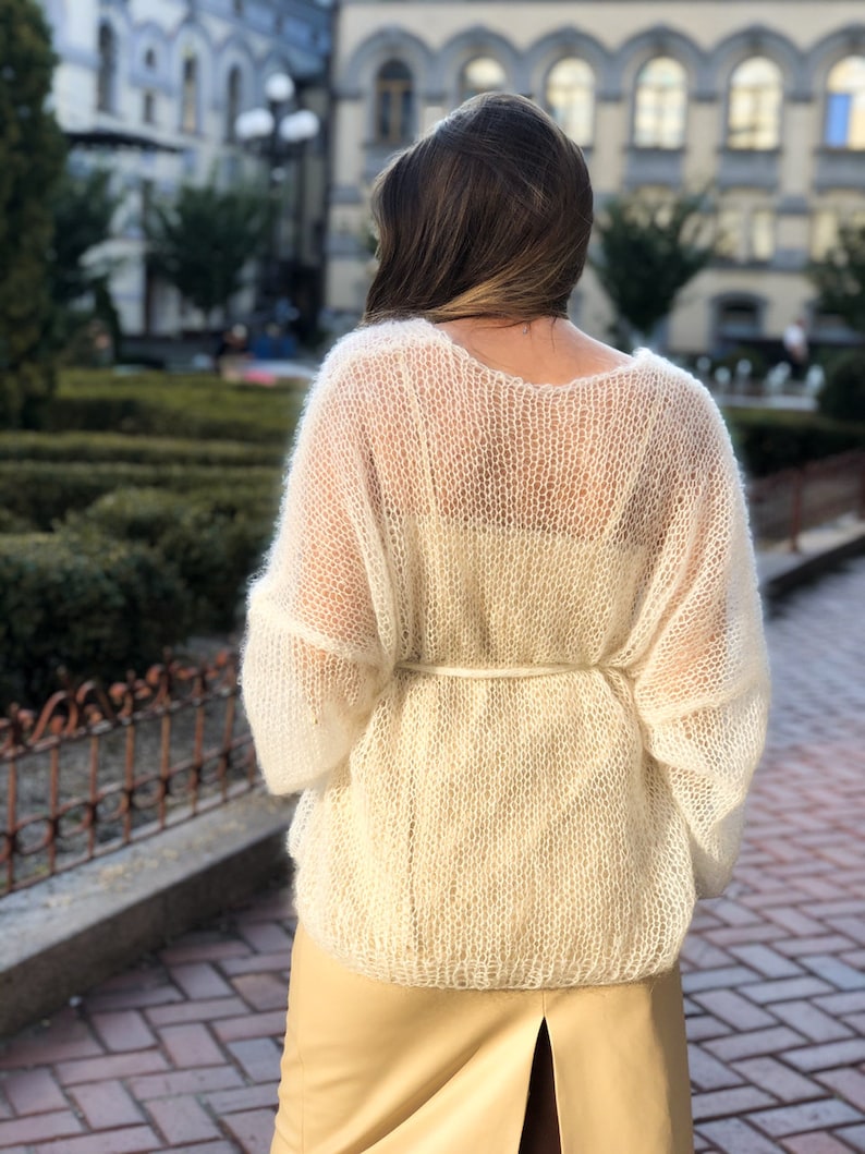 May include: A white, sheer, knitted sweater with a loose fit and long sleeves. The sweater is tied at the waist with a white ribbon.