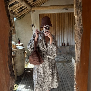 Puede incluir: Una mujer con un vestido estampado en marrón y blanco, un bolso de crochet marrón y un sombrero marrón. La mujer está de pie en una habitación con paredes y suelos de madera.