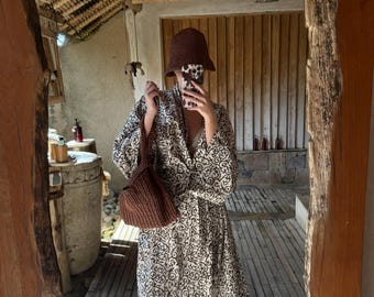 May include: A woman wearing a brown and white patterned dress with a brown crocheted bag and a brown hat. The woman is standing in a room with wooden walls and floors.