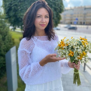 May include: A woman in a white, lacy, knitted sweater holds a bouquet of yellow and white flowers. She is wearing a white skirt and has long brown hair.