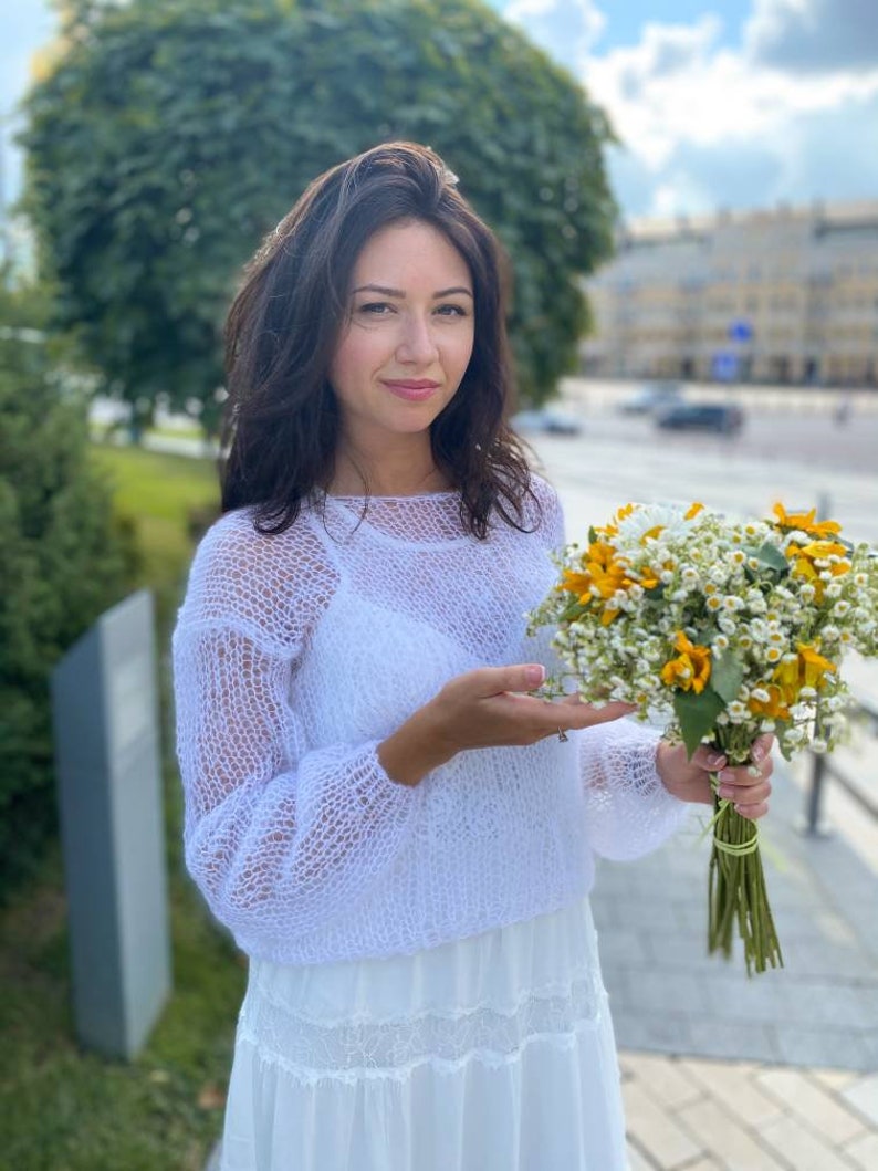 May include: A woman in a white, lacy, knitted sweater holds a bouquet of yellow and white flowers. She is wearing a white skirt and has long brown hair.