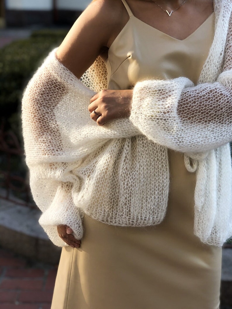 May include: A white, open-knit cardigan with a delicate, lacy pattern. The cardigan is worn over a beige slip dress.