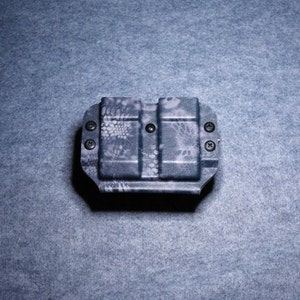 May include: A black and gray patterned magazine carrier with four screws. The carrier has a camouflage-like design and is made of a durable material. The carrier is designed to hold two magazines.
