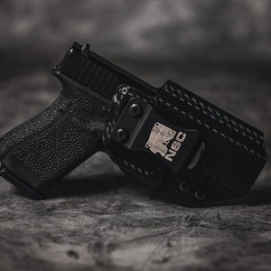 May include: A black handgun with a black carbon fiber holster. The holster has a metal clip with the text "NSC" on it.
