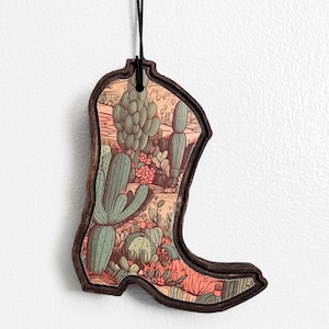 May include: Wooden cowboy boot-shaped ornament with a desert landscape design. The interior features a detailed illustration of cacti, succulents, and desert plants in shades of green, pink, and brown. The boot has a dark brown border and a black string for hanging.