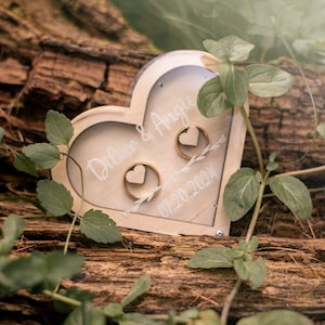 Może przedstawiać: Drewniane pudełko na obrączki w kształcie serca z przezroczystą pokrywką, z napisem "Dillon & Angie 07.20.2024". Wewnątrz pudełka znajdują się dwie złote obrączki.