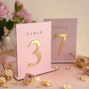 Puede incluir: Dos tarjetas de número de mesa de color rosa con números dorados. La tarjeta de la izquierda tiene el número 3 y la tarjeta de la derecha tiene el número 7. Ambas tarjetas tienen la palabra "TABLE" impresa en ellas.