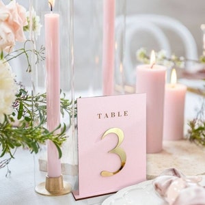 May include: A wedding table setting with pink candles, floral arrangements, and a pink table number card with a gold number 3. The candles are lit, and the table is set for a celebration.