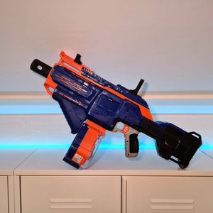 Könnte beinhalten: Ein blauer und orangefarbener Nerf-Blaster mit einem schwarzen Lauf und einem Magazin. Der Blaster hat eine taktische Schiene oben und einen Griff unten.