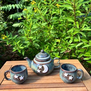 May include: A ceramic tea set with a teal glaze, featuring a teapot and two mugs. Each piece is adorned with white crescent moons and stars. The set is displayed on a wooden table, with lush green foliage in the background.