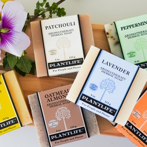May include: Assortment of Plantlife aromatherapy herbal soaps, including Patchouli, Peppermint, Lavender, and Vanilla Orange. Each bar is 4 oz (113g) and features a unique label. A purple and white flower and greenery are in the background.