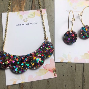 以下が含まれることがあります： 虹色のグリッターが散りばめられた、波打つような襟と丸い形の虹色のグリッターのイヤリングが付いた、金のネックレスとイヤリングのセット。ネックレスとイヤリングは、「ARS STUDIO 74」と書かれた白いカードの上に置かれています。