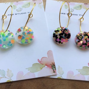Puede incluir: Dos pares de pendientes de aro dorados con dijes redondos de resina transparente. Un par tiene un diseño de confeti con purpurina rosa, azul, verde y naranja. El otro par tiene un diseño de confeti con purpurina negra, rosa y dorada. Los pendientes se muestran en tarjetas blancas con diseños florales.