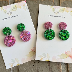 May include: Two pairs of glitter dangle earrings. Each earring has a small green glitter circle above a larger pink glitter circle. The earrings are displayed on white cards with the text "ARS STUDIO 74".