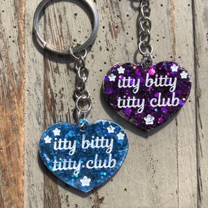 Op de afbeelding: Twee hartvormige sleutelhangers met glitter ingebed in de hars. De sleutelhangers zijn blauw en paars en hebben de tekst "itty bitty titty club" in wit.
