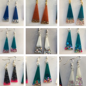 Op de afbeelding: Een set van tien paar driehoekige oorbellen van hars met glitter ingebed in de hars. De oorbellen zijn in verschillende kleuren, waaronder turquoise, blauw, wit, zwart en oranje.