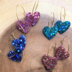 Op de afbeelding: Set van zes gouden creolen met hartvormige bedels. De bedels zijn gevuld met iriserende glitter in tinten roze, blauw en turquoise.