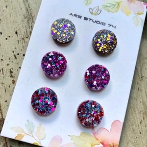 May include: Six round, flat, glitter-covered, resin buttons in various colours, including silver, gold, pink, and blue. The buttons are arranged in two rows of three on a white card with the text "ARS STUDIO 74" printed on it.