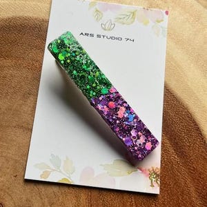 Peut inclure: Une barrette rectangulaire avec un dégradé de paillettes vertes à violettes. La barrette est posée sur une carte blanche avec le texte "ARS STUDIO 74" et des motifs floraux. La barrette est en résine.