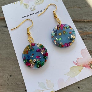 Puede incluir: Un par de pendientes de resina azul con purpurina dorada y multicolor incrustada en la resina. Los pendientes están en una tarjeta blanca con el texto "ARS STUDIO 74".