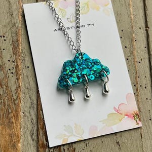 May include: A silver chain necklace with a teal cloud pendant filled with glitter and three silver raindrop charms. The pendant is on a white card with the text "ARE STUDIO 74".