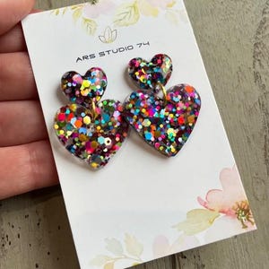 May include: Glitter heart earrings with a smaller heart on top. The earrings are multi-colored with a variety of glitter colors. The earrings are on a white card with the text "ARS STUDIO 74".